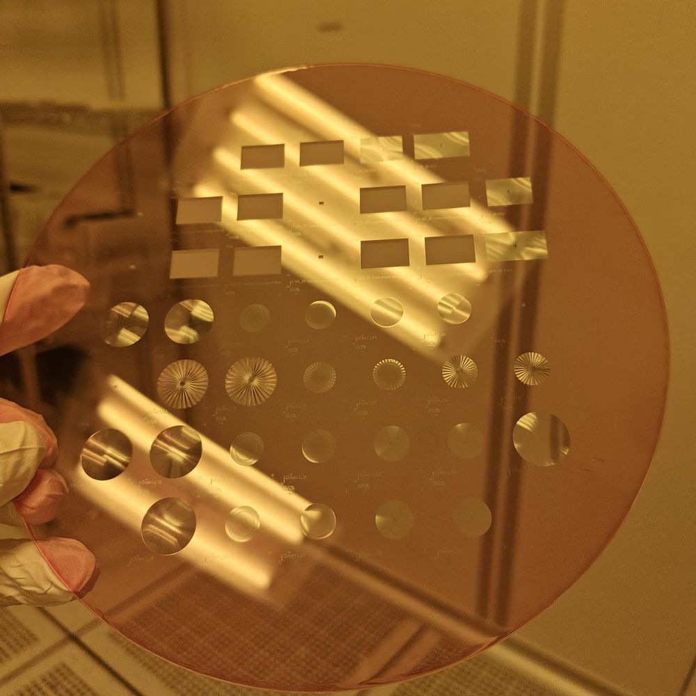 A wafer of flat lenses and holograms held up in the cleanroom's yellow light.